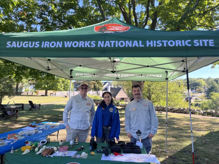 Three interns outdoors behind a table with accessible birding materials, under a tent that says Saugus Iron Works National Historic Site.