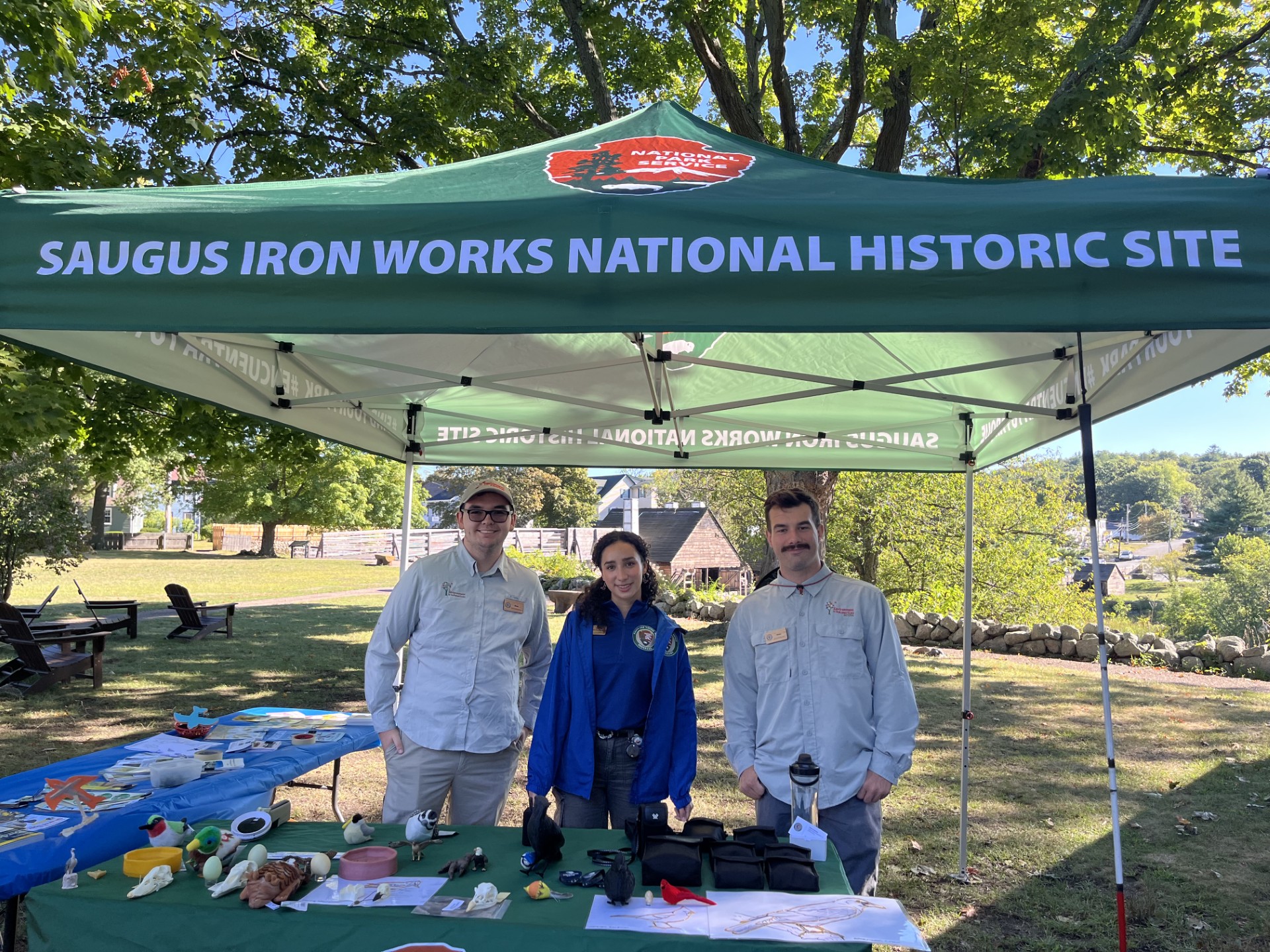 Three interns outdoors behind a table with accessible birding materials, under a tent that says Saugus Iron Works National Historic Site.